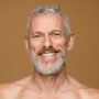 Smiling Caucasian middle-aged shirtless man with grey hair beard isolated. Male beauty concept Smiling with toothy smile caucasian mature middle-aged shirtless man with grey hair and beard looking at camera isolated in dark beige background. Male beauty concept